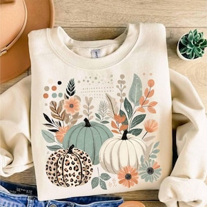 May include: A cream-coloured sweatshirt with a graphic print of three pumpkins surrounded by green and brown leaves and flowers. The pumpkins are green, white, and brown with a leopard print.