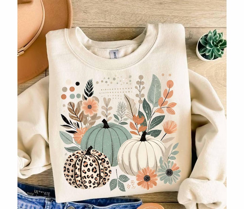 May include: A cream-coloured sweatshirt with a graphic print of three pumpkins surrounded by green and brown leaves and flowers. The pumpkins are green, white, and brown with a leopard print.