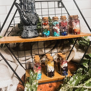 May include: A black metal shelf with two wooden shelves displays small glass bottles filled with colorful gemstones. A black hand statue with intricate designs sits on the top shelf. The bottles are capped with corks, and the background is a white tiled wall.