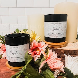 May include: Two black soy wax candles with wooden lids. The candles are labeled "Imbolc" and feature the text "Lore of the Pomegranates". The candles are on a wooden surface with pink flowers.