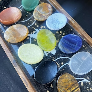May include: A collection of oval-shaped polished stones in various colors, including green, brown, white, yellow, blue, black, and orange, arranged in a wooden tray with a black background.