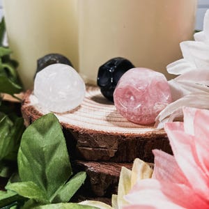 May include: Four small crystal skull figurines on a wooden surface. The skulls are white, black, pink, and gray. The skulls are surrounded by green leaves and pink flowers.