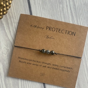 May include: A black string bracelet with four obsidian beads. The card reads "the little band of PROTECTION - Obsidian" and "Promotes protection, strength, clarity, compassion. Boosts your sense of self and shields negativity."