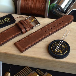 Könnte beinhalten: Braunes Lederarmband mit silberner Schnalle, präsentiert auf einer Holzoberfläche. Das Armband hat detaillierte Nähte und wird mit einem schwarzen Etikett mit goldenem Text, Nadeln und einem kleinen runden Emblem gezeigt.