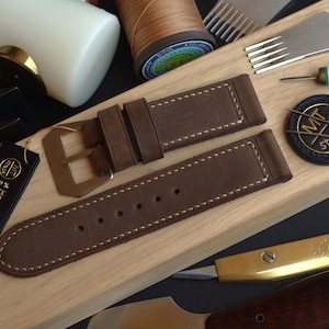 Puede incluir: Una correa de reloj de cuero marrón con una hebilla dorada, con costuras blancas. La correa se muestra con herramientas de marroquinería, incluyendo un mazo, agujas y una bobina de hilo. Una etiqueta dice "Hecho a mano en Italia".