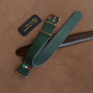 May include: A green leather watch strap with a gold buckle and a small Italian flag on the strap. The strap is on a wooden surface.