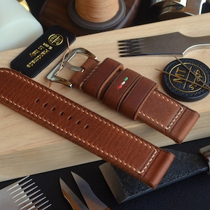 Può includere: Un cinturino per orologio in pelle marrone con cuciture bianche e fibbia argentata. Il cinturino presenta un dettaglio della bandiera italiana ed è esposto con strumenti di pelletteria e un'etichetta nera con scritto "Handmade in Italy".