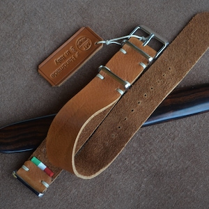May include: A brown leather watch strap with white stitching and a silver buckle. The strap has a small Italian flag on it and a tag that says "Handmade in Italy".