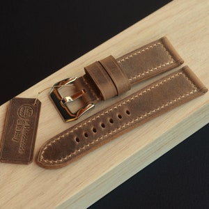 May include: A brown leather watch strap with white stitching and a gold buckle. The strap is 100% handmade in Italy.
