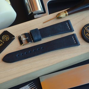 Pode incluir: Close-up de pulseiras de relógio em couro azul marinho com costuras brancas e fivelas prateadas, juntamente com ferramentas de artesanato em couro. Uma etiqueta preta com texto dourado diz "Handmade in Italy".