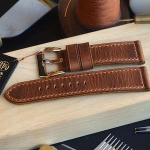 Peut inclure: Un bracelet de montre en cuir marron avec une boucle argentée, posé sur une surface en bois. Le bracelet présente des coutures et une texture vieillie. Une étiquette indique "Handmade in Italy". D'autres outils et matériaux sont en arrière-plan.