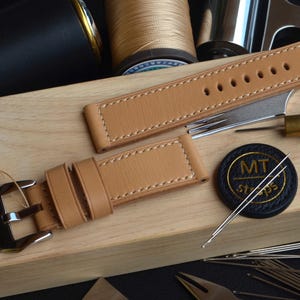 Puede incluir: Correa de reloj de cuero color canela con costuras blancas y hebilla plateada. Se encuentra sobre una superficie de madera con herramientas de relojero, carretes de hilo y una etiqueta negra que dice "100% Handmade in Italy".