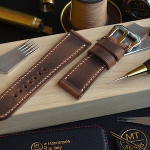 May include: Brown leather watch strap with white stitching, a silver buckle, and a set of watchmaking tools. The strap is on a wooden block, with other tools and materials in the background. The tag reads "Handmade in Italy."
