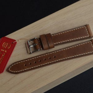 Può includere: Cinturino per orologio in pelle marrone con cuciture bianche e fibbia argentata. Un'etichetta rossa con testo dorato è allegata. Il cinturino è esposto su una superficie di legno chiaro.