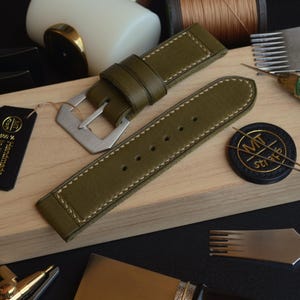 Puede incluir: Correa de reloj de cuero verde oliva con costuras blancas y hebilla plateada. La correa se muestra sobre una superficie de madera junto con herramientas de relojería y una etiqueta negra con texto dorado "100% Handmade in Italy."