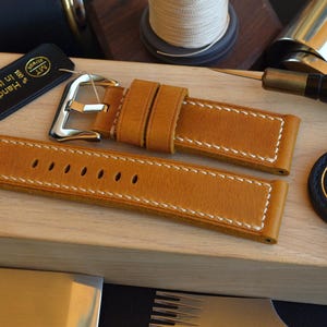 May include: A tan leather watch strap featuring white contrast stitching and a polished silver-tone metal buckle. The strap includes two leather keepers and a series of oval adjustment holes. A black tag labelled MT straps Handmade in Italy is included.