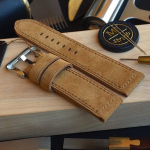 May include: A tan suede watch strap with a silver buckle, presented on a wooden surface. A black tag with gold text reading "100% Handmade in Italy" is attached. Watchmaking tools, including a needle, are also visible.