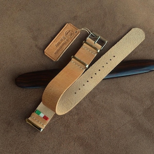 Può includere: Un cinturino per orologio in pelle color cuoio chiaro con fibbia e passanti in metallo argentato. Il cinturino presenta una piccola etichetta con la bandiera italiana e un cartellino rettangolare in pelle con il testo Handmade in Italy. Un oggetto in legno scuro si trova sotto.