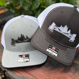 May include: Two black and grey trucker hats with white mesh panels. Both hats feature a white embroidered design of two axes crossed with a forest scene in between. The hats have a black and white label with the text "Richardson" and "Original 112 Trucker Adjustable".