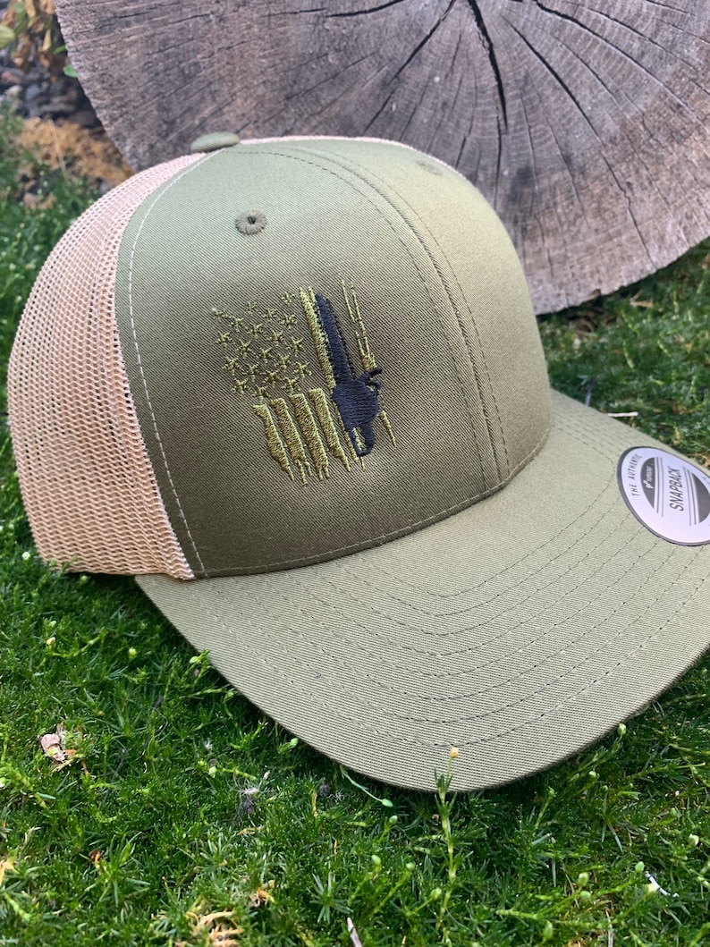 May include: A green and tan trucker hat with a distressed American flag design and a black silhouette of an eagle. The hat has a mesh back and a curved brim.