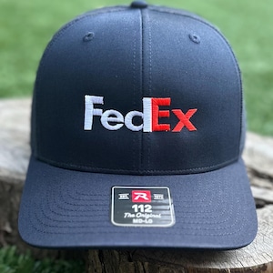 May include: A navy blue baseball cap featuring the white and red FedEx logo embroidered on the front. A black sticker on the brim displays the text "EST. 1970", "R", "112", "The Original", and "MD-LG" in white and red lettering.
