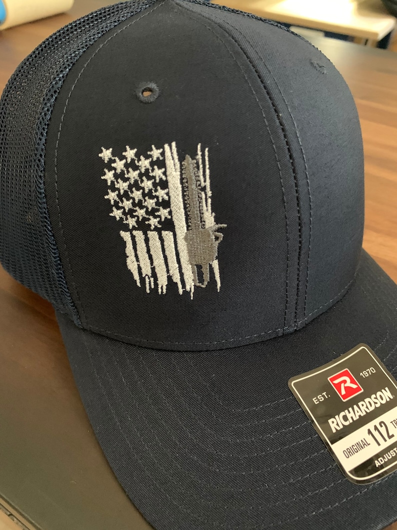 May include: A navy blue baseball cap with a black mesh back. The front of the cap features an embroidered American flag with a faded, distressed look. The cap has a black adjustable strap with a white label that reads "EST. 1970 RICHARDSON ORIGINAL 112 ADJUSTABLE".