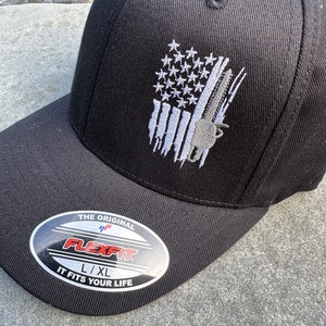 May include: Black baseball cap with a white embroidered American flag design and a gray chainsaw graphic. The cap has a Flexfit label with the text "The Original Flexfit L/XL It Fits Your Life".