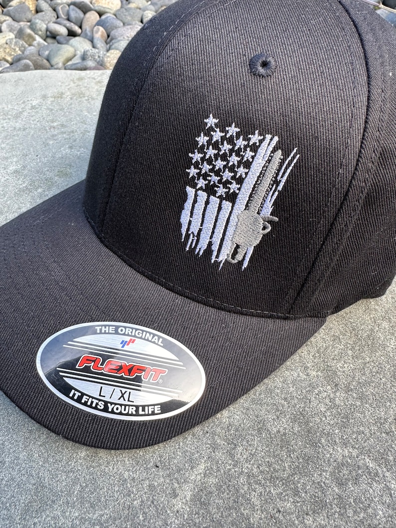May include: Black baseball cap with a white embroidered American flag design and a gray chainsaw graphic. The cap has a Flexfit label with the text "The Original Flexfit L/XL It Fits Your Life".