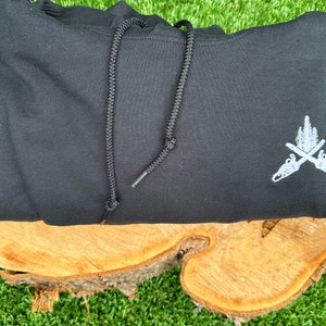 May include: Black hooded sweatshirt with a white embroidered design of two crossed axes with a tree on top.