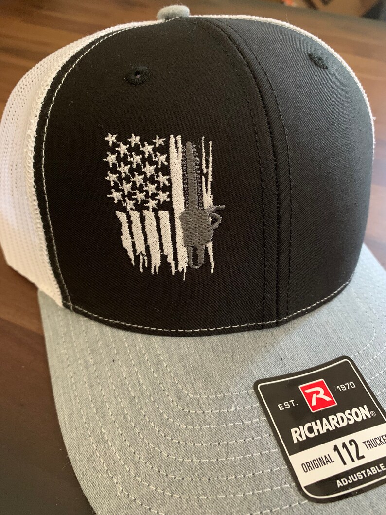 May include: Black and white trucker hat with a distressed American flag design and a gray underbill. The hat features a gray silhouette of a deer in the flag design. The hat is adjustable and has a Richardson 112 label with the text "EST. 1970 RICHARDSON ORIGINAL TRUCKER ADJUSTABLE".