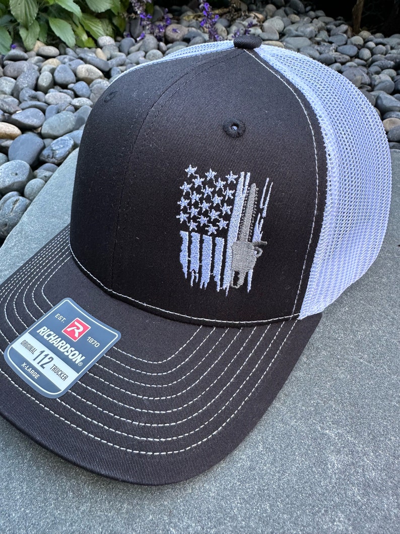 May include: Black and white trucker hat with a white mesh back. The front of the hat features an embroidered American flag with a rifle and a hunting dog. The hat has a Richardson 112 label with the text "Original 112 Trucker" and "X-Large".