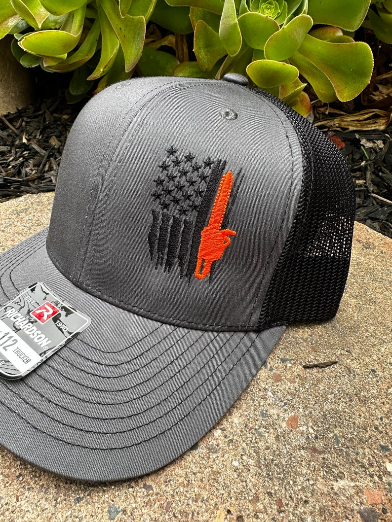 May include: A gray and black trucker hat with an embroidered American flag design and a chainsaw. The flag is black and white with an orange chainsaw.