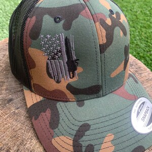 May include: A camouflage trucker hat with a black mesh back and a gray embroidered American flag with a silhouette of a bald eagle. The hat has a black snap closure and a "The Authentic Yupoong Snapback" label.