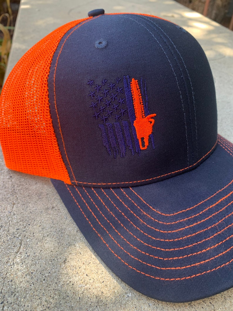 May include: A navy blue and orange trucker hat with a purple and orange embroidered American flag design featuring a chainsaw. The hat has a mesh back and a curved brim.