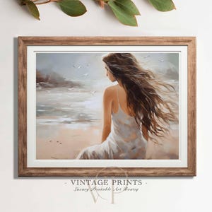 Puede incluir: Una impresión artística enmarcada representa a una mujer sentada en una playa, frente al mar. Lleva un vestido blanco y su largo cabello castaño ondea al viento. La obra utiliza tonos suaves y apagados, con gaviotas en el fondo. El marco es de color marrón cálido.
