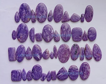 May include: A collection of polished purple charoite gemstones in various shapes including ovals, teardrops, and rectangles. Each stone features swirling white and dark violet patterns. The text UGJEWELLERS is visible on several stones.