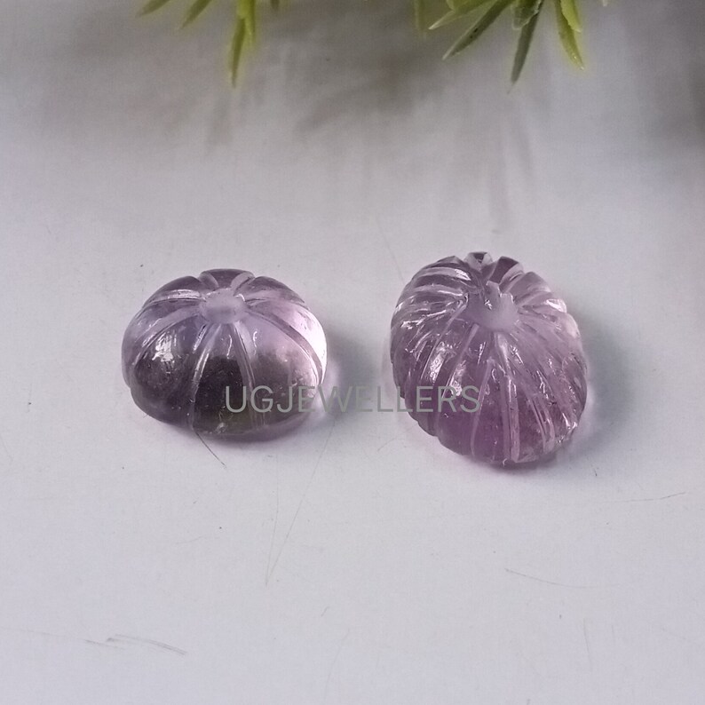 K&ouml;nnte beinhalten: Zwei geschnitzte Amethyst-Edelsteine, jeweils in K&uuml;rbisform. Die Edelsteine sind helllila und haben detaillierte geschnitzte Linien. Die Edelsteine liegen auf einer wei&szlig;en Oberfl&auml;che.