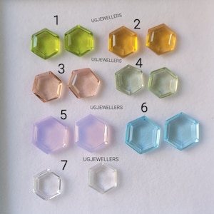 Op de afbeelding: Zeven zeshoekige edelstenen in verschillende kleuren, waaronder groen, geel, roze, lavendel, blauw en helder. Elke edelsteen is genummerd van 1 tot 7. De tekst "UGJEWELLERS" staat drie keer op de afbeelding.