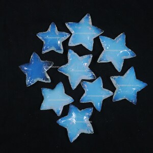 Puede incluir: Nueve piedras de opalita azul en forma de estrella con un brillo iridiscente blanco. Las piedras están dispuestas en semicírculo sobre un fondo negro. El texto "UNITEDGEMSJWELLERS" está impreso en cada piedra.