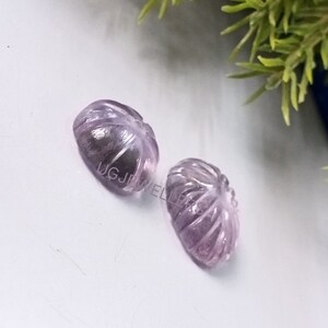 K&ouml;nnte beinhalten: Zwei ovale, geschnitzte Edelsteine in Lavendel- und Lilat&ouml;nen. Die Edelsteine sind mit einem blattartigen Muster mit detaillierten Adern versehen. Sie liegen auf einer wei&szlig;en Oberfl&auml;che, mit einem Hauch von Gr&uuml;n im Hintergrund.