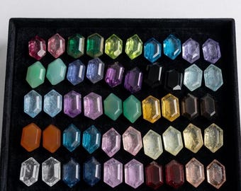 May include: A collection of numerous hexagonal-cut gemstones arranged in a black display tray. The stones feature a wide variety of colors including red, pink, green, blue, purple, black, yellow, orange, and clear, all with faceted surfaces.