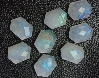 May include: Seven iridescent white moonstone cabochons, each cut into a hexagonal shape, are arranged on a black textured surface. The stones have a subtle blue and green sheen.