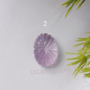 K&ouml;nnte beinhalten: Ein geschnitzter, oval geformter, lavendelfarbener Edelstein mit radialen Rillen und einem zentralen Loch. Die Zahl "2" befindet sich &uuml;ber dem Edelstein. Der Text "UGJEWELLERS" ist darunter gedruckt. Gr&uuml;nes Laub befindet sich im Hintergrund.