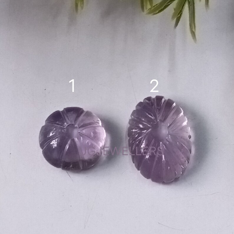 K&ouml;nnte beinhalten: Zwei geschnitzte Amethyst-Edelsteine. Einer ist rund, &auml;hnelt einem K&uuml;rbis, der andere ist oval. Beide haben detaillierte, geschnitzte Rippen, die von einem zentralen Punkt ausgehen. Die Edelsteine haben eine durchscheinende violette Farbe.