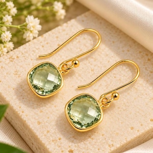 May include: A pair of gold drop earrings featuring square-cut, faceted light green gemstones. Each earring is suspended from a simple gold hook wire, showcasing a polished metal setting that frames the translucent, pale green stones.
