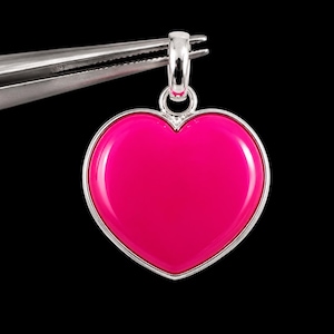 May include: A heart-shaped pendant featuring a smooth, vibrant hot pink center encased in a polished silver-toned metal frame. The pendant includes a small, circular silver-toned bail at the top, designed for attachment to a necklace chain.