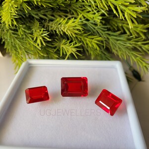 Könnte beinhalten: Drei rote rechteckige Cabochons aus Edelstein in einer weißen Schachtel. Die Edelsteine sind unterschiedlich groß. Die Schachtel trägt den Schriftzug "UGJEWELLERS" auf der Unterseite.