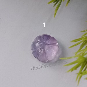 K&ouml;nnte beinhalten: Ein geschnitzter, hellvioletter Edelstein, m&ouml;glicherweise Amethyst, in Form einer Blume oder eines K&uuml;rbisses. Der Edelstein hat detaillierte geschnitzte Linien und ist mit der Nummer "1" versehen. Der Text "UG JEWELLERS" ist sichtbar.