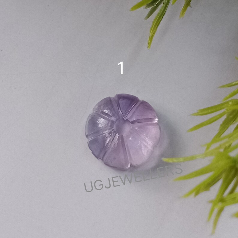 K&ouml;nnte beinhalten: Ein geschnitzter, hellvioletter Edelstein, m&ouml;glicherweise Amethyst, in Form einer Blume oder eines K&uuml;rbisses. Der Edelstein hat detaillierte geschnitzte Linien und ist mit der Nummer "1" versehen. Der Text "UG JEWELLERS" ist sichtbar.