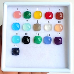 May include: A white tray containing 16 different coloured gemstone cabochons, numbered 1 to 16. The cabochons are arranged in four rows of four. The tray is on a white surface.